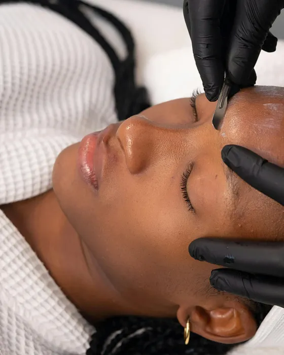 A dermaplaning blade being drawn across a client’s forehead at a shallow angle while she lies back with her eyes closed
