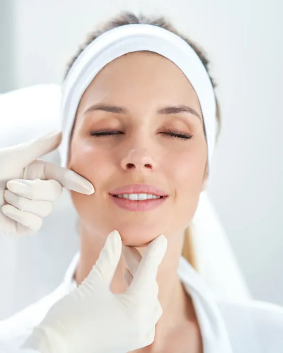 A close crop of a face during a treatment, with a headband holding the hair back and a gloved hand resting at the cheek