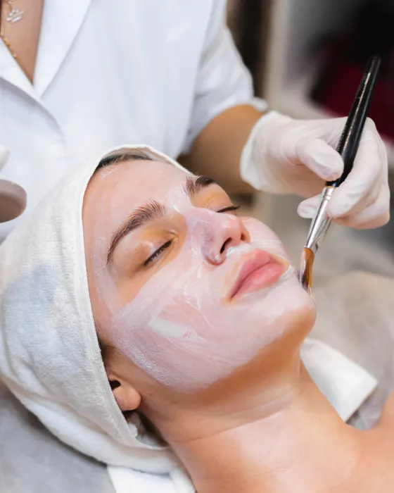 A gloved hand brushing a peel solution across the face of a client lying back with a white headband on and eyes closed